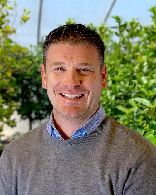 A man in a gray sweater standing in front of plants.