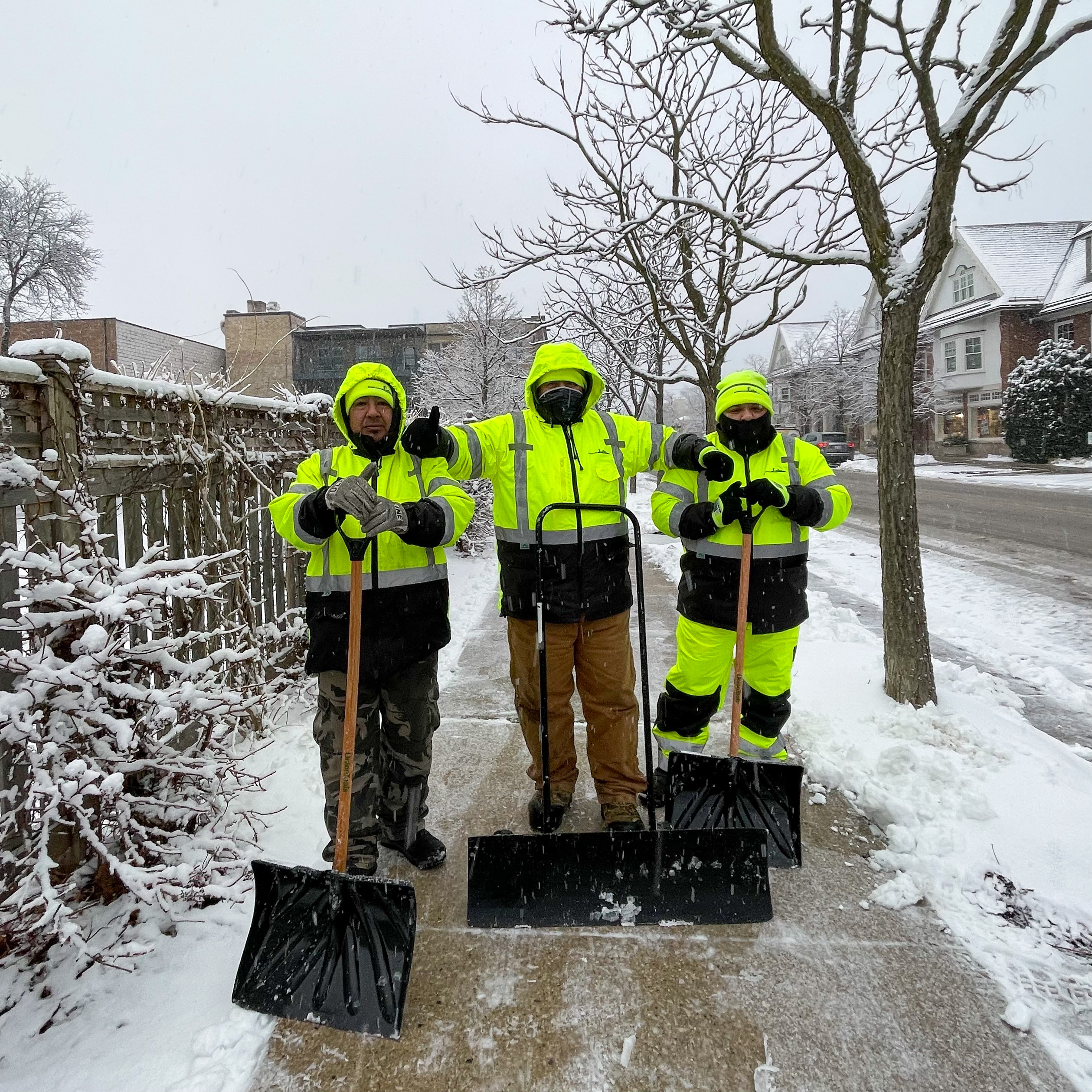 Three individuals in high-visibility jackets holding shovels on a snowy residential street.