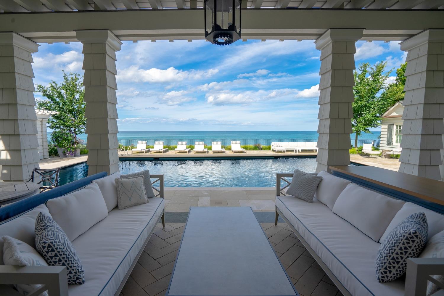 View from a covered patio with two couches overlooking a swimming pool and a calm sea under a clear sky.