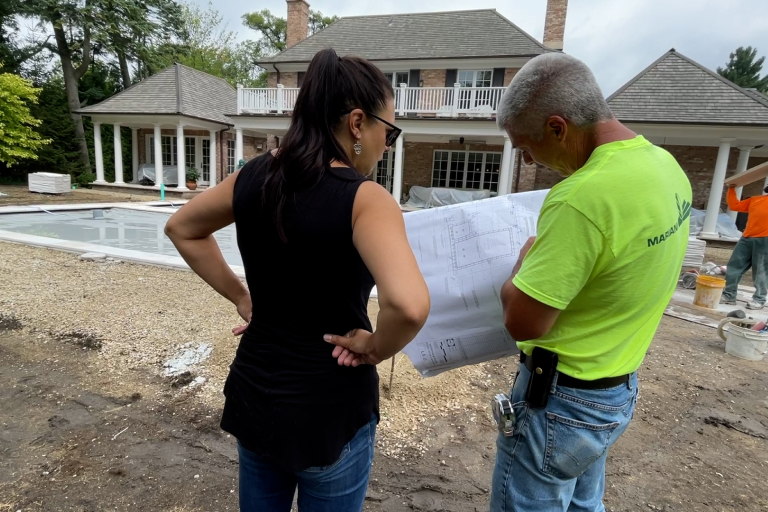 Two people, a woman and a man, review blueprints at a residential construction site with a partially completed house and pool in the background.