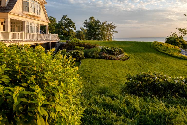 A large house with a porch overlooking a manicured lawn and a distant ocean view under a clear, sunny sky.