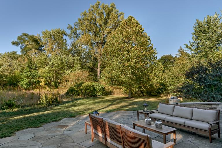 A serene backyard featuring a stone patio with a sofa and chairs, surrounded by lush trees and a neatly trimmed lawn.