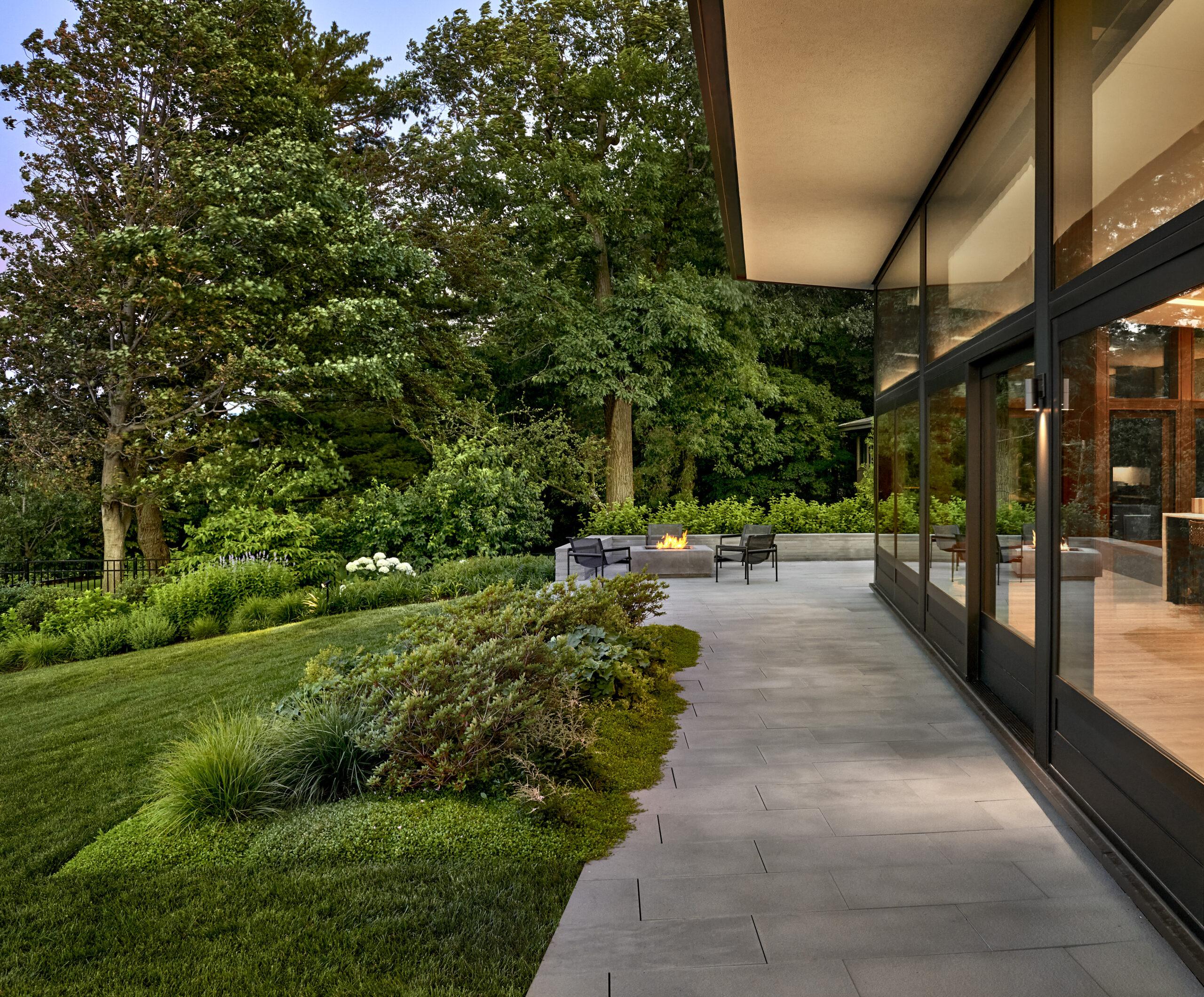 Modern house with large glass windows overlooking a landscaped patio with outdoor seating, surrounded by lush green lawn and trees.