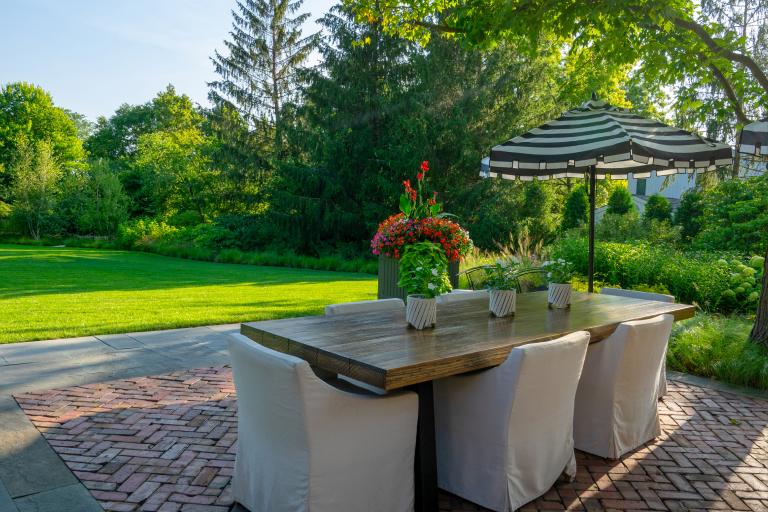 Outdoor dining setup with a wooden table and chairs under a striped umbrella, surrounded by lush greenery and a neatly manicured lawn.