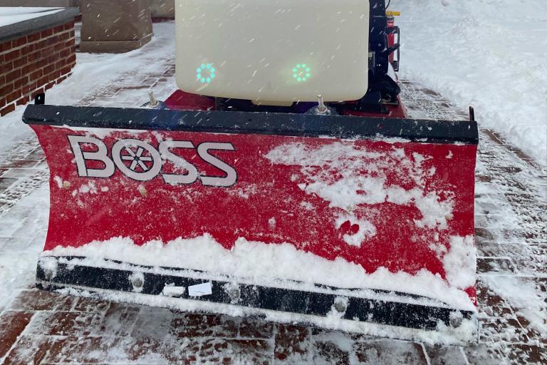 A snow-covered "boss" brand plow attached to a vehicle, clearing snow, with light flurries and a brick structure in the background.