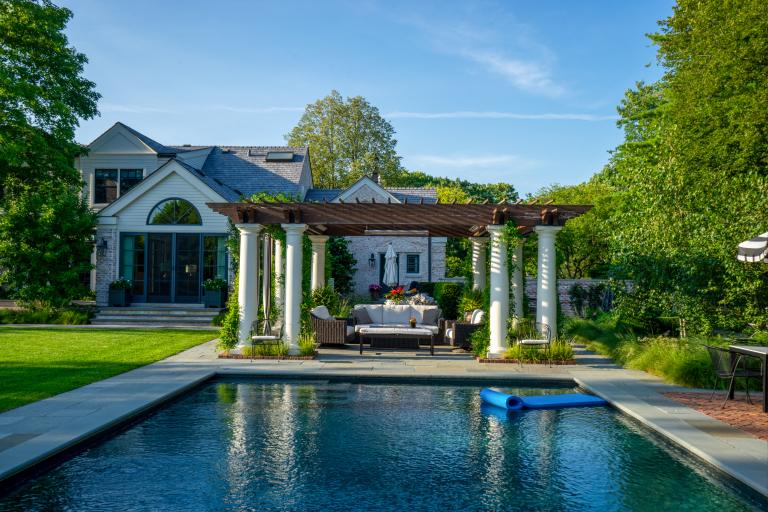 Luxurious backyard with a swimming pool, pergola with sitting area, and a large house surrounded by lush greenery.