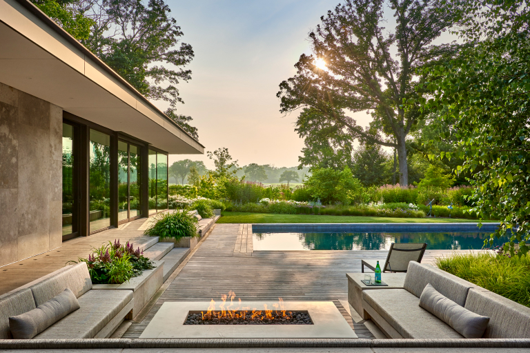 Modern backyard with a rectangular pool, outdoor fire pit, seating area, and a minimalist house with large glass windows overlooking a serene lake.