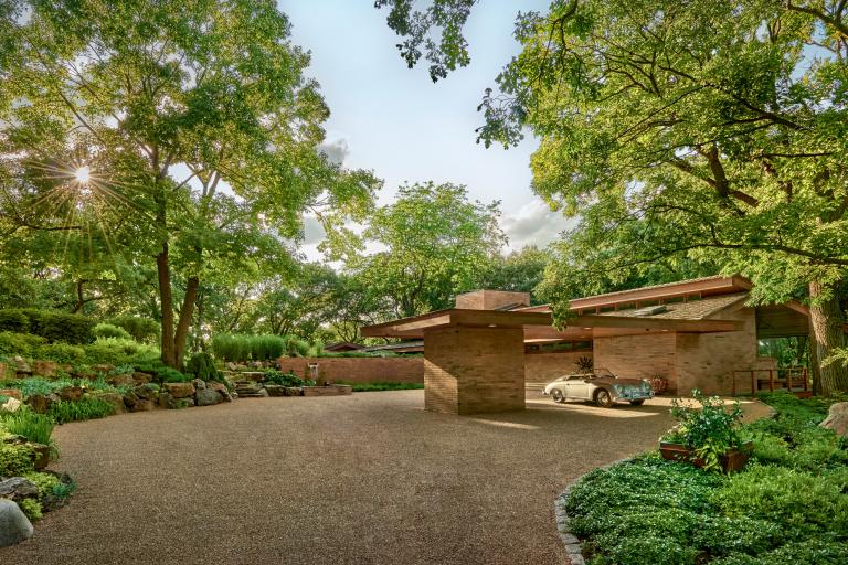 A vintage car parked in front of a frank lloyd wright-style house amidst lush greenery and trees, with sunlight peeking through foliage.