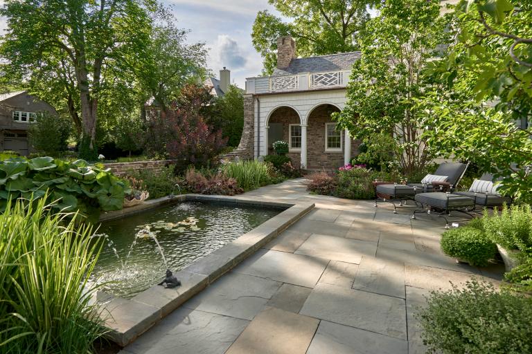 A serene backyard garden with a rectangular pond, surrounded by lush greenery and a pathway leading to an elegant house with a white balcony.