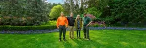Four people stand on a well-manicured lawn with a garden of trees and flowers in the background.