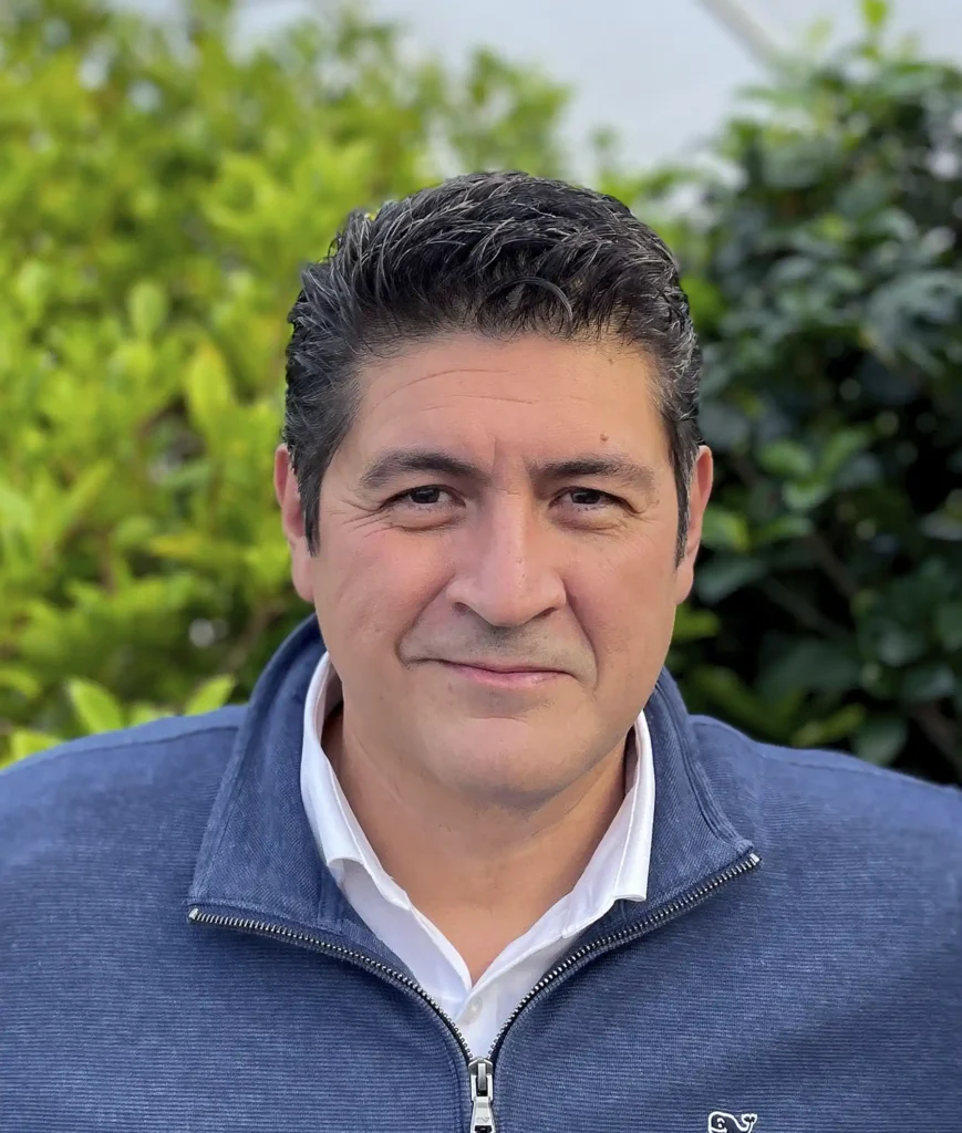 Man with short dark hair wearing a white collared shirt and blue zip-up jacket, standing in front of green foliage.