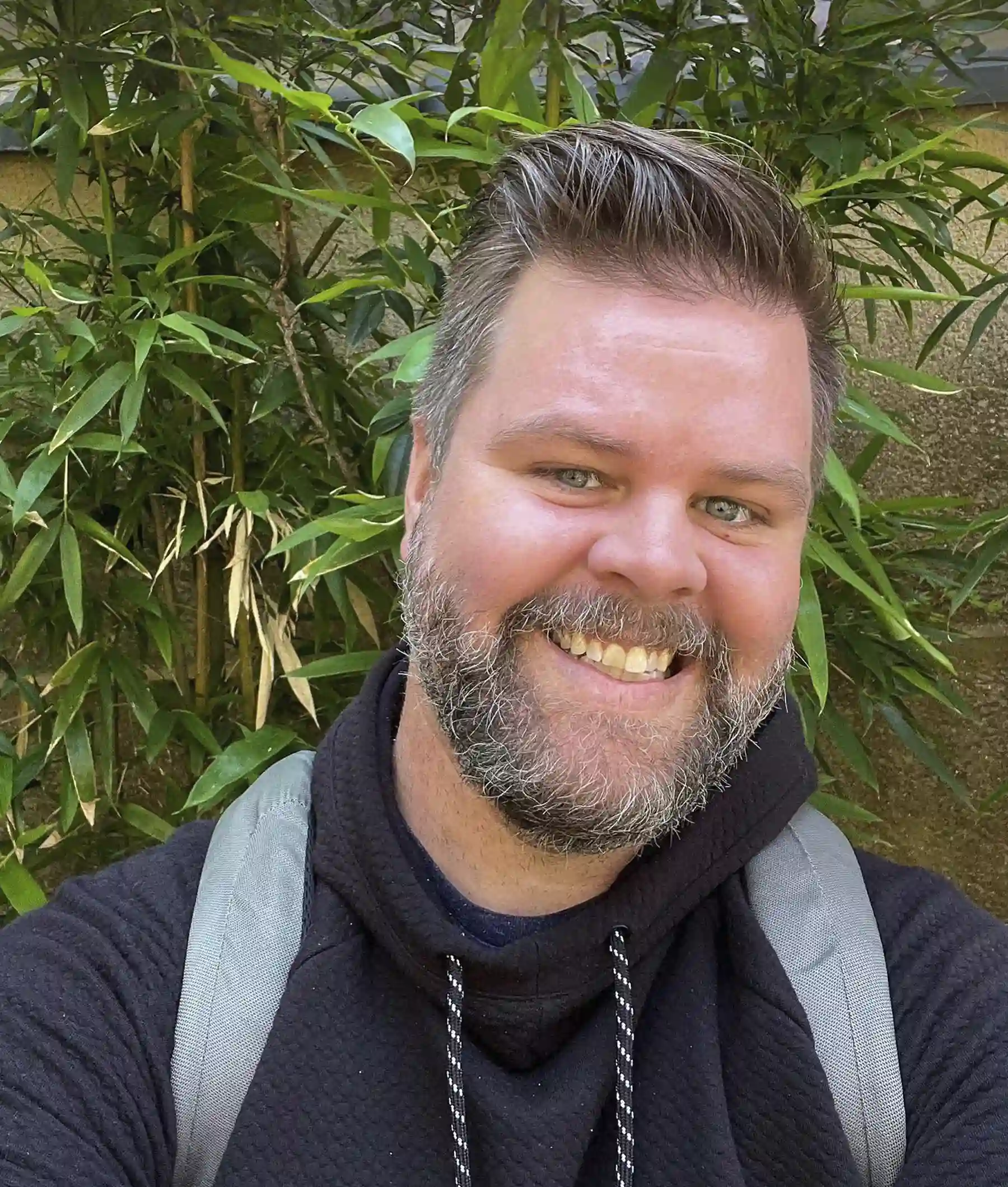 A man with a beard and backpack smiles in front of green foliage. He is wearing a black hoodie.