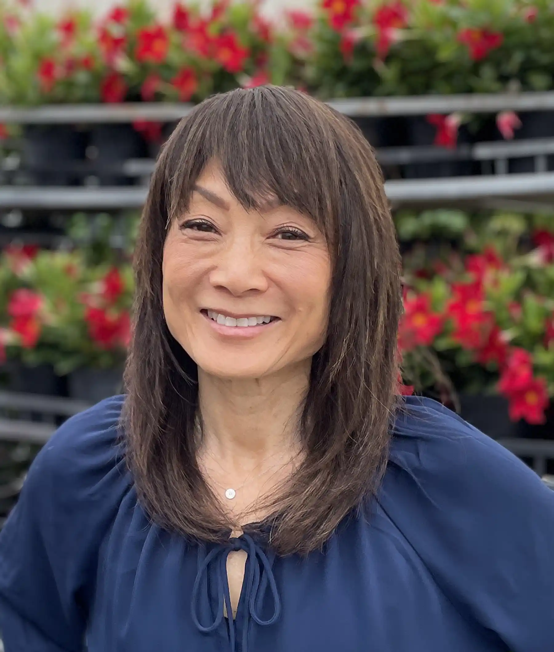 A woman with straight brown hair wearing a blue top smiles in front of a background with red flowers and potted plants.