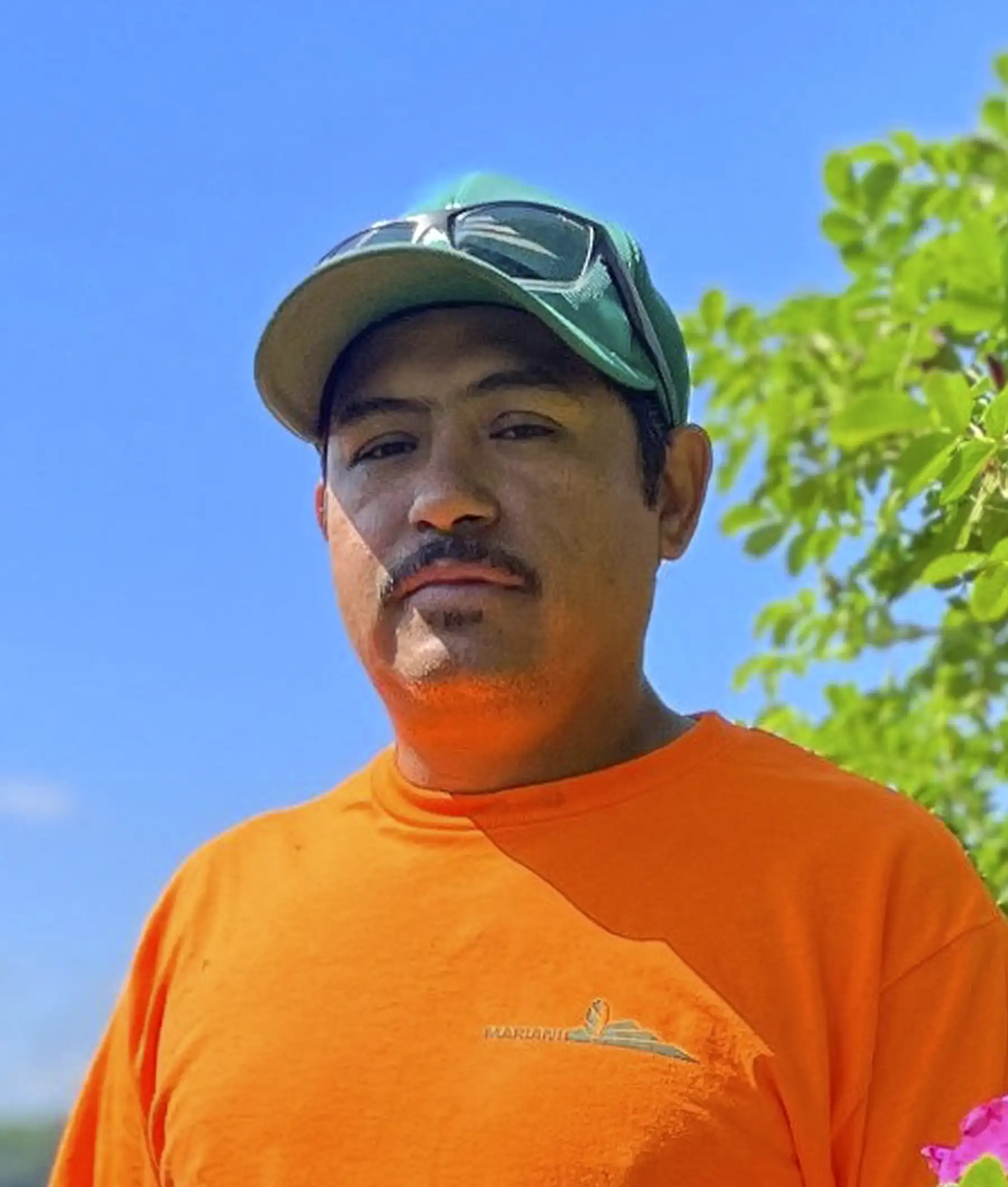 A man wearing an orange shirt and green cap stands outdoors with greenery in the background.