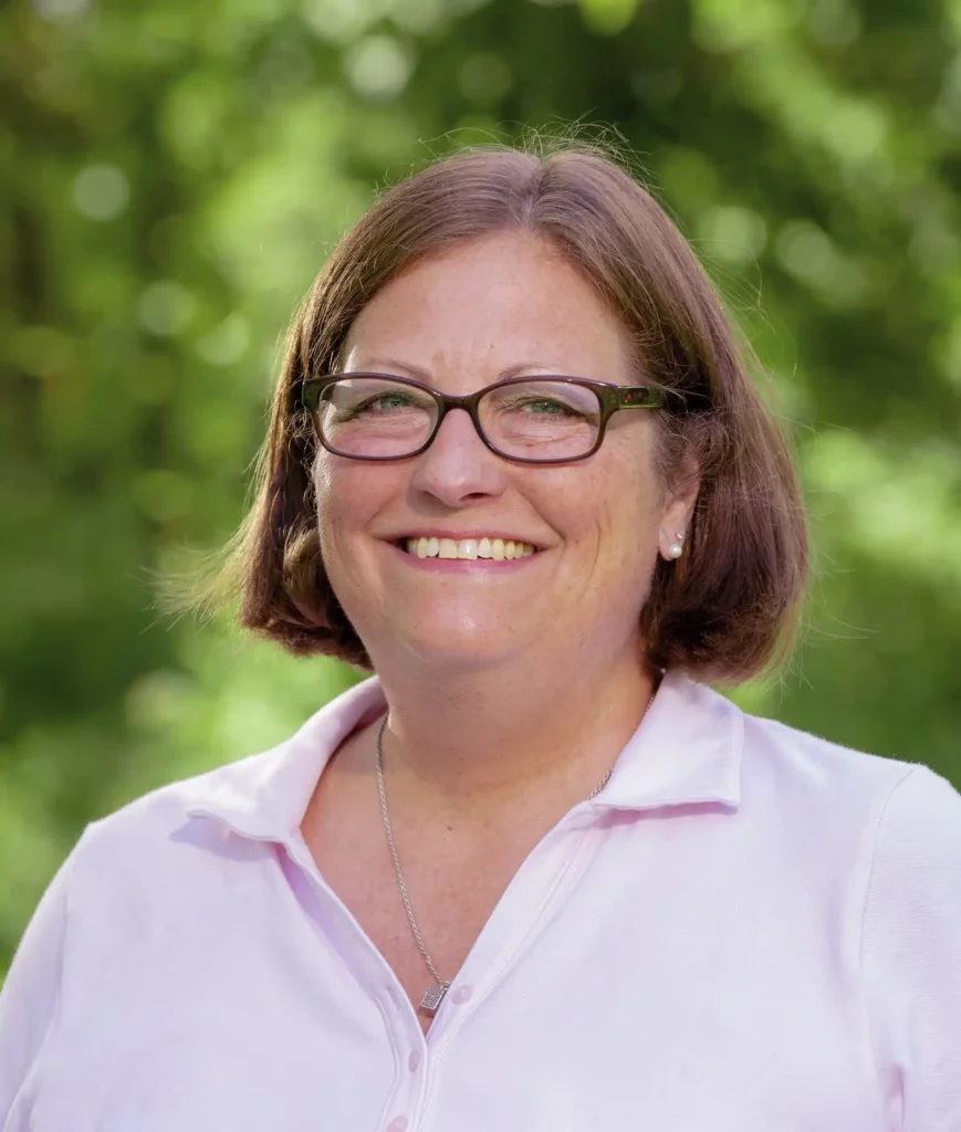A woman with glasses and short brown hair smiles while wearing a light pink polo shirt, standing in front of a green, leafy background.