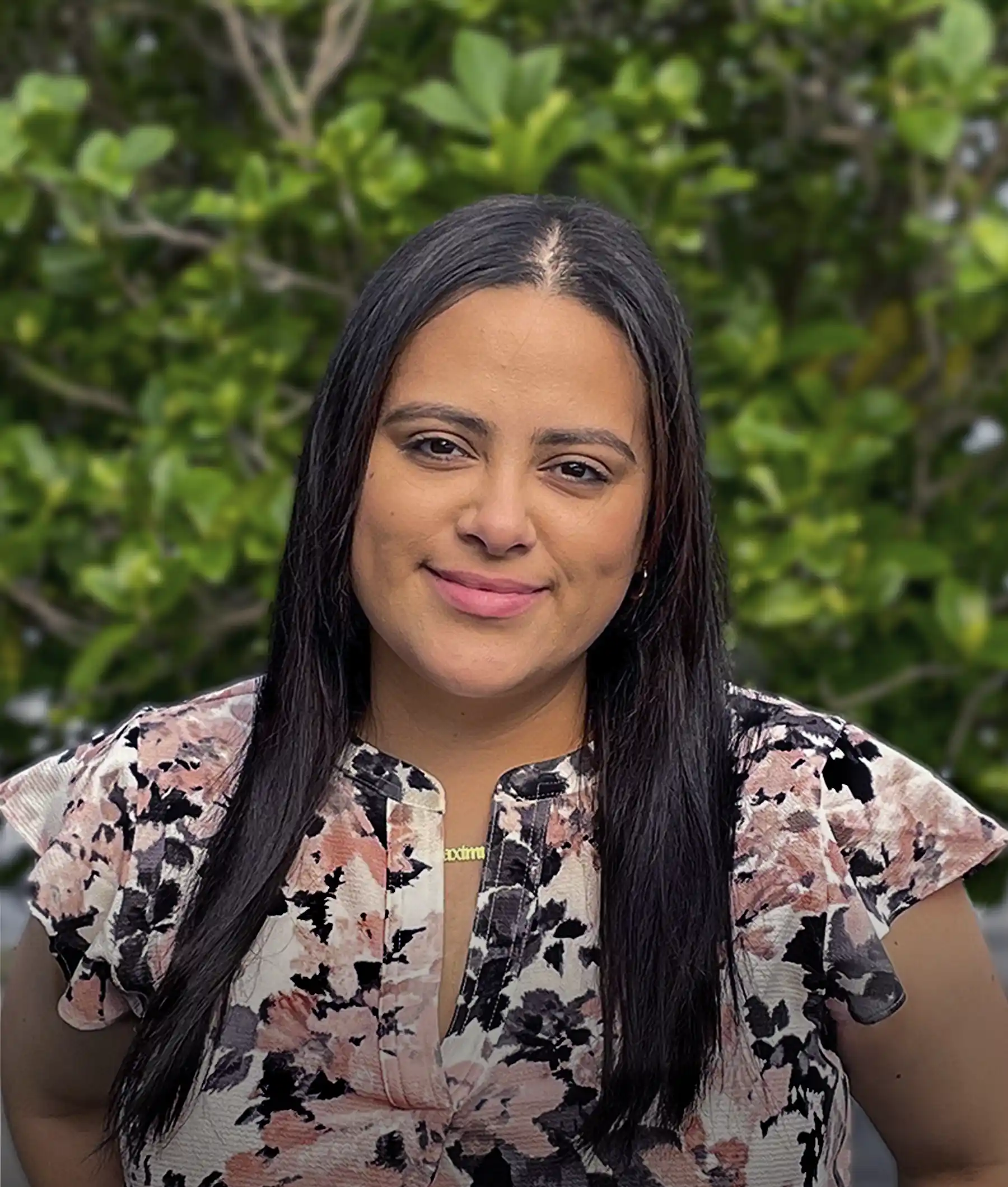 A person with long dark hair and a floral blouse stands in front of a green leafy background, smiling at the camera.
