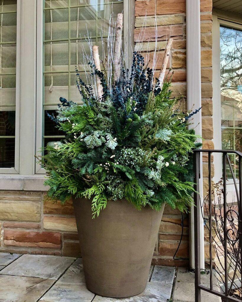 Large pot filled with a mix of evergreen branches, birch branches, and dark berried stems placed on a stone surface next to a house window with a stone exterior.