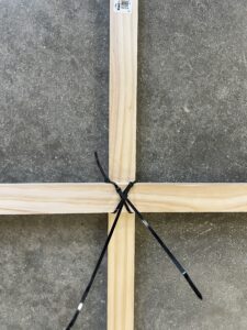 A wooden frame with two black zip ties securing the intersecting beams on a gray stone floor.
