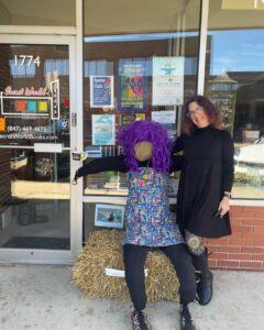A woman stands smiling next to a purple-haired scarecrow sitting on a hay bale outside a bookstore.