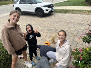 Three people are posing outdoors. A child is making a playful gesture. A pumpkin is placed next to a stuffed scarecrow on the ground. A white SUV is parked on the street.