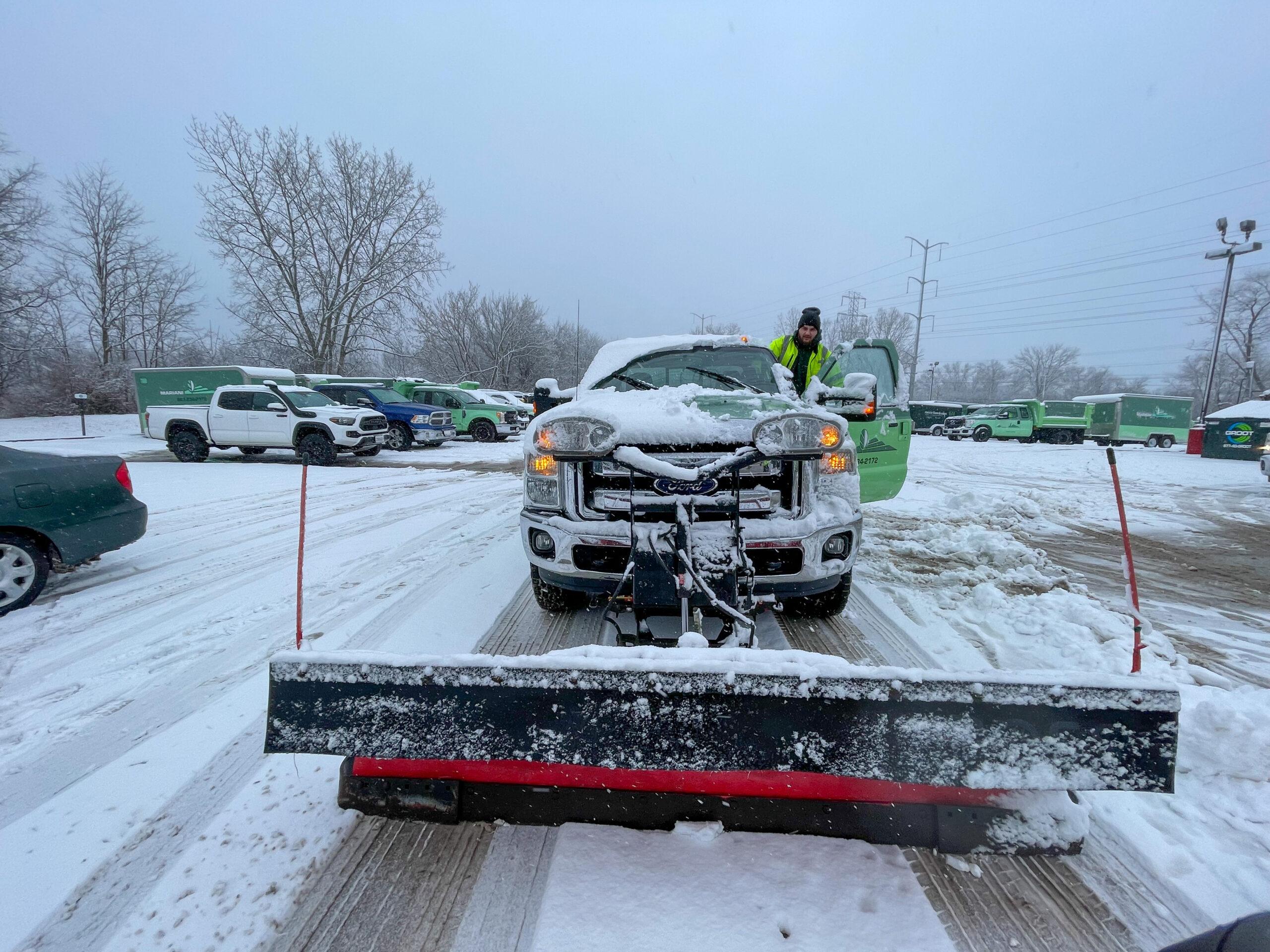 A snow plow truck with snow on its hood is parked on a snowy lot, expertly prepared for Snow & Ice Removal for HOAs. A person in winter gear exits the driver's side door. Several vehicles and trees create a frosty backdrop.