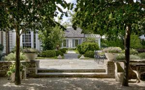 A garden with stone steps leading to a patio, surrounded by lush greenery and large potted plants. A table and chairs are set on the right side under trees.