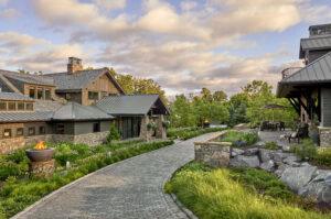 Large stone house with multiple roofs and windows, surrounded by lush greenery and a stone pathway. A patio with furniture is visible on the right, set against a backdrop of trees and cloudy skies.