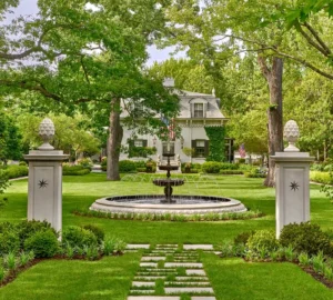 A stately house with a manicured garden, featuring a central circular fountain, stone path, and gate pillars topped with decorative stone pineapples.