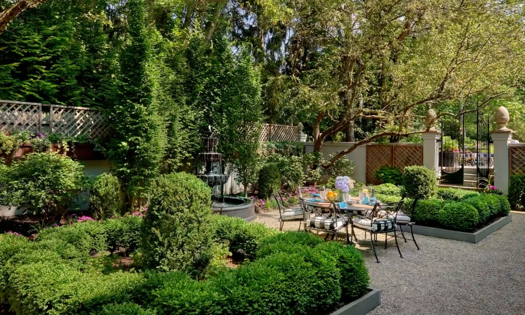 A garden with a gravel path, manicured bushes, patio furniture, a dining table with decor, and a tiered fountain surrounded by trees and lush greenery.