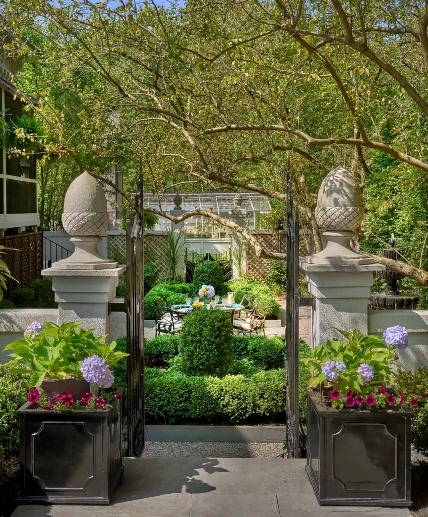 Elegant garden with stone pillars and iron gates, featuring lush greenery, potted flowers, and a patio set with a table and chairs under a tree canopy.