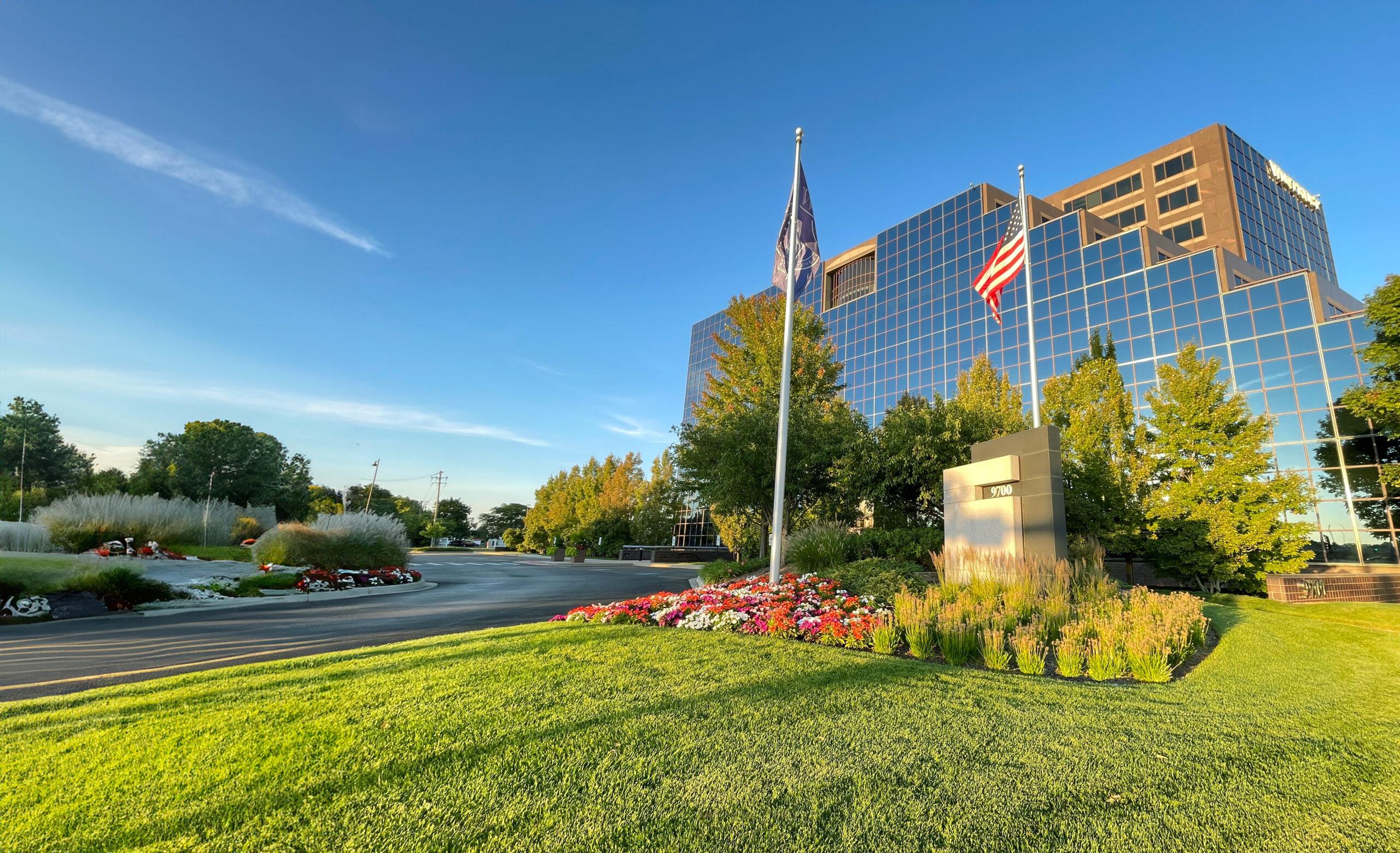 A modern glass building with multiple floors stands behind a meticulously maintained landscape area. Expert landscape maintenance for business parks ensures vibrant flowers, flags, and lush greenery flourish under a clear blue sky.