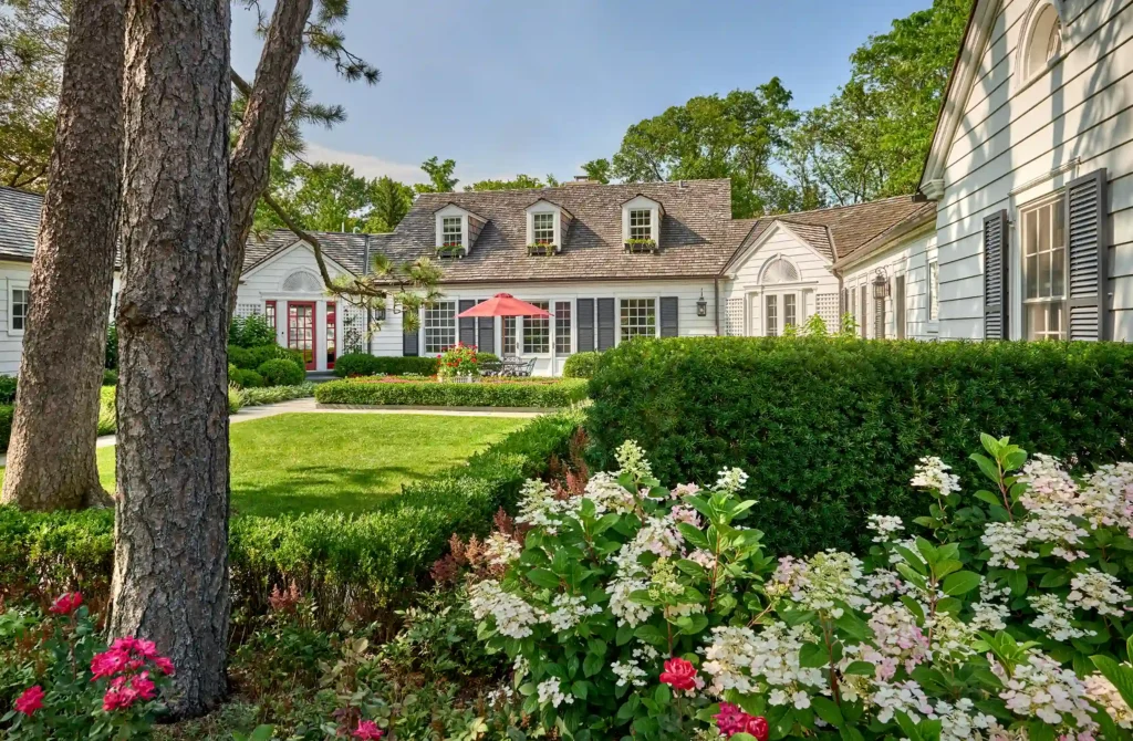 Charming white house with gray shutters, surrounded by lush greenery and flowers. Manicured lawn, red patio umbrella, and clear blue sky.