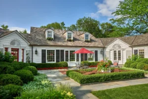 White colonial-style house with black shutters, surrounded by a lush garden. A red umbrella shades patio furniture. Blue sky and trees in the background.