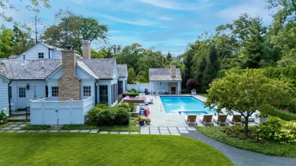 Aerial view of a house with a chimney, adjacent patio, and a swimming pool surrounded by trees and lounge chairs on a paved area. Lawn and path in the foreground. Clear blue sky overhead.