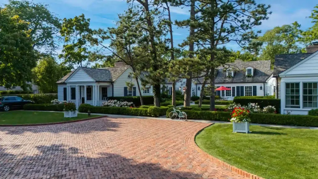 A brick driveway leads to a large white house surrounded by trees and landscaped gardens. A bicycle is parked in front, and a red umbrella is visible in the backyard.