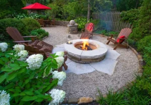 A garden with a gravel patio features wooden chairs around a stone fire pit. A table with a red umbrella is in the background, surrounded by greenery and white flowers.
