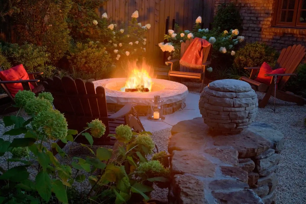 Outdoor seating area with wooden chairs around a lit fire pit. Lanterns and plants surround the scene, creating a cozy atmosphere.