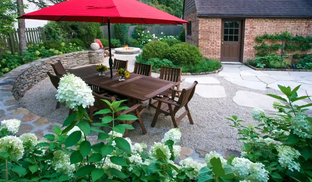 Patio with a wooden dining set under a red umbrella, surrounded by blooming hydrangeas. Stone path leads to a brick building with a dark wooden door.