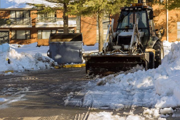 A snowplow efficiently tackles snow and ice removal for HOAs, clearing a parking lot near a brick apartment building on a sunny winter day.