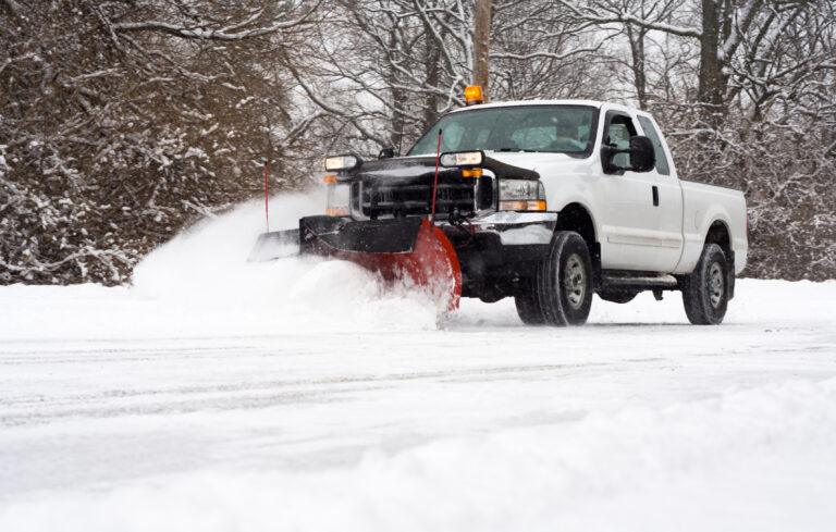 A white truck with a snowplow attachment efficiently handles snow and ice removal for hospitals, ensuring clear pathways. In the background, snow-covered trees create a serene winter landscape.