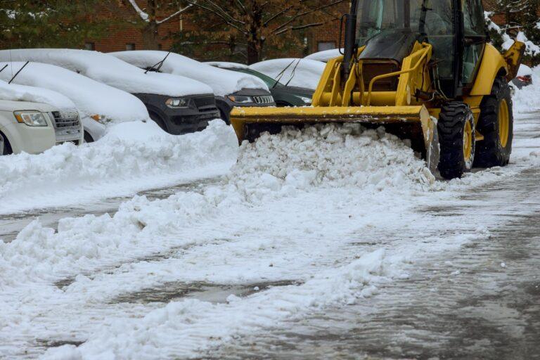 A yellow snowplow clears snow from a parking lot lined with snow-covered cars, providing efficient snow and ice removal for HOAs.