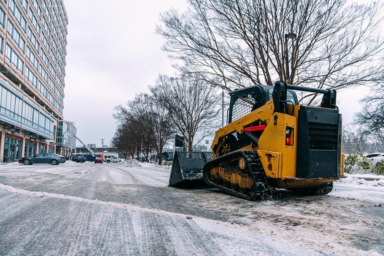 A yellow skid-steer loader, essential for Snow & Ice Removal for HOAs, is parked on a snowy urban street. It's flanked by bare trees and modern buildings in the background.