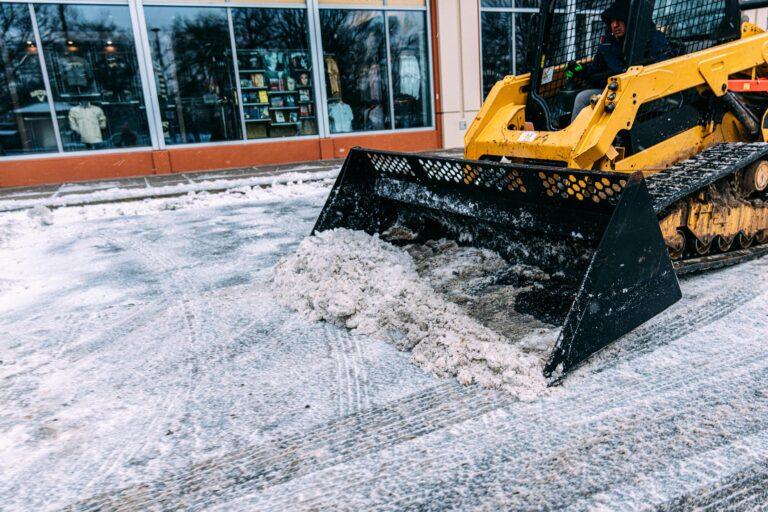 A yellow skid steer loader clears snow from a pavement in front of store windows.