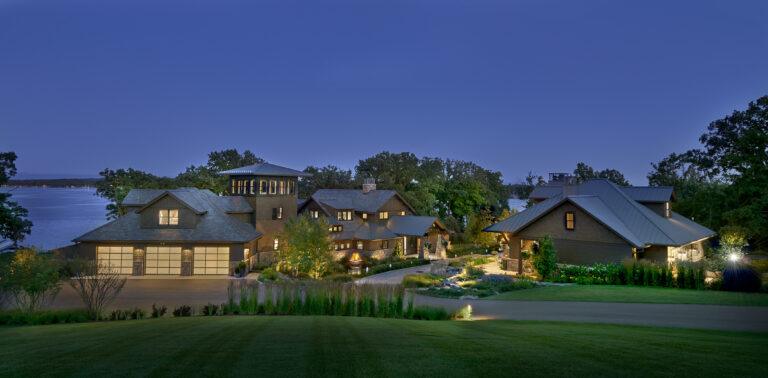 Large modern lakeside home complex with multiple buildings, surrounded by trees and landscaping, illuminated at dusk with a view of the water in the background.