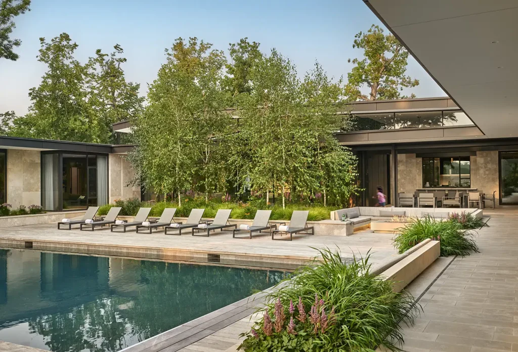 Modern backyard with a large swimming pool, several lounge chairs on a stone deck, lush greenery, and a contemporary house in the background.