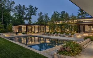Modern house exterior at dusk with large windows, a rectangular swimming pool, poolside lounge chairs, and landscaped greenery.