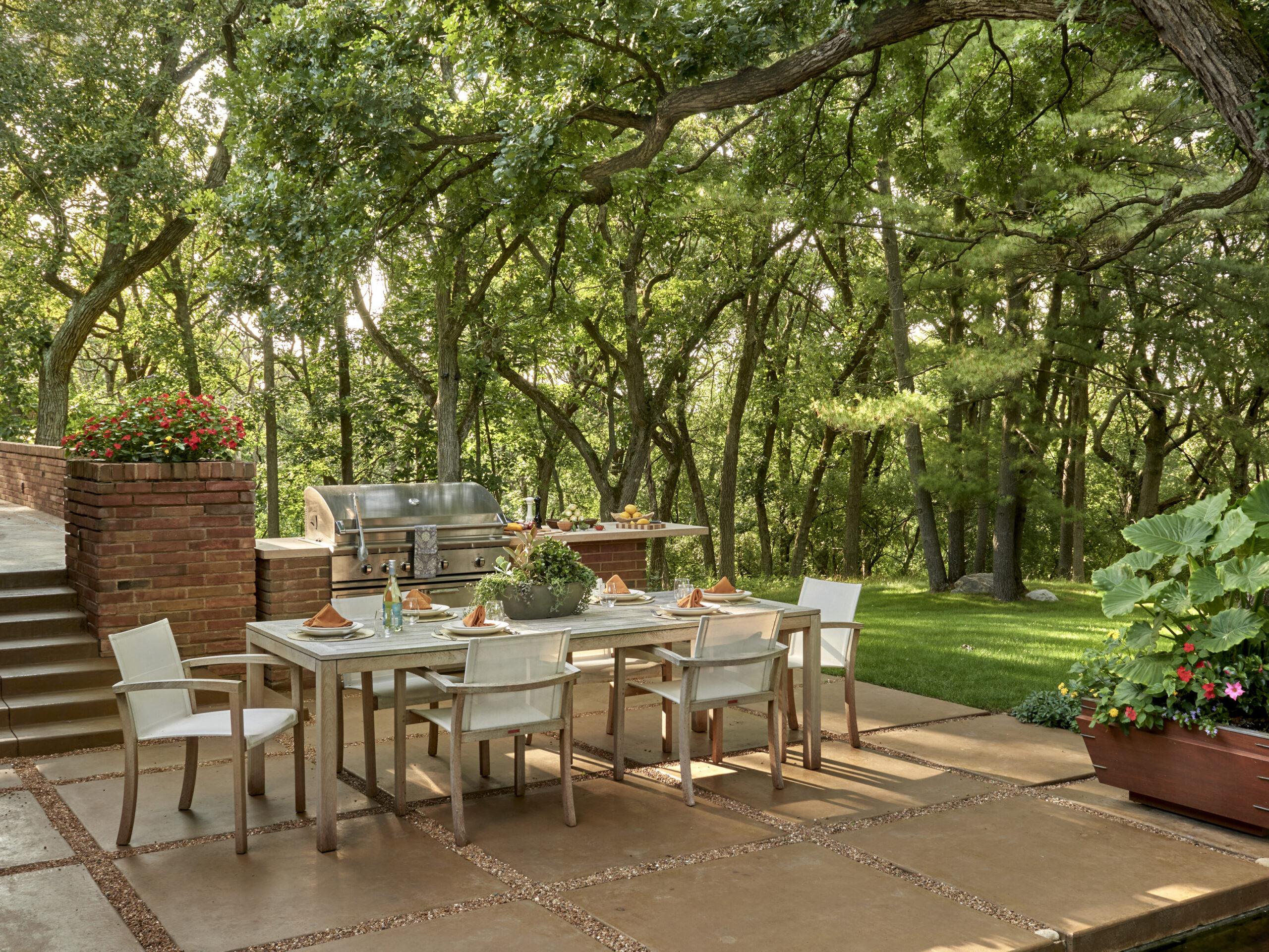 A modern outdoor dining area with a glass table set for six, a stainless steel grill, potted plants, and lush greenery—perfect for enjoying expertly designed landscaping in Barrington IL.