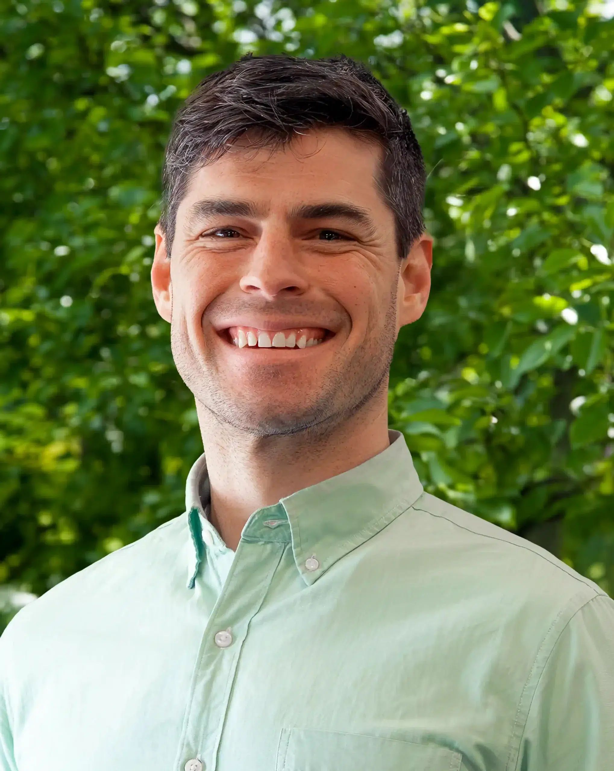 A man wearing a light green button-up shirt smiles in front of a background of green leafy trees.