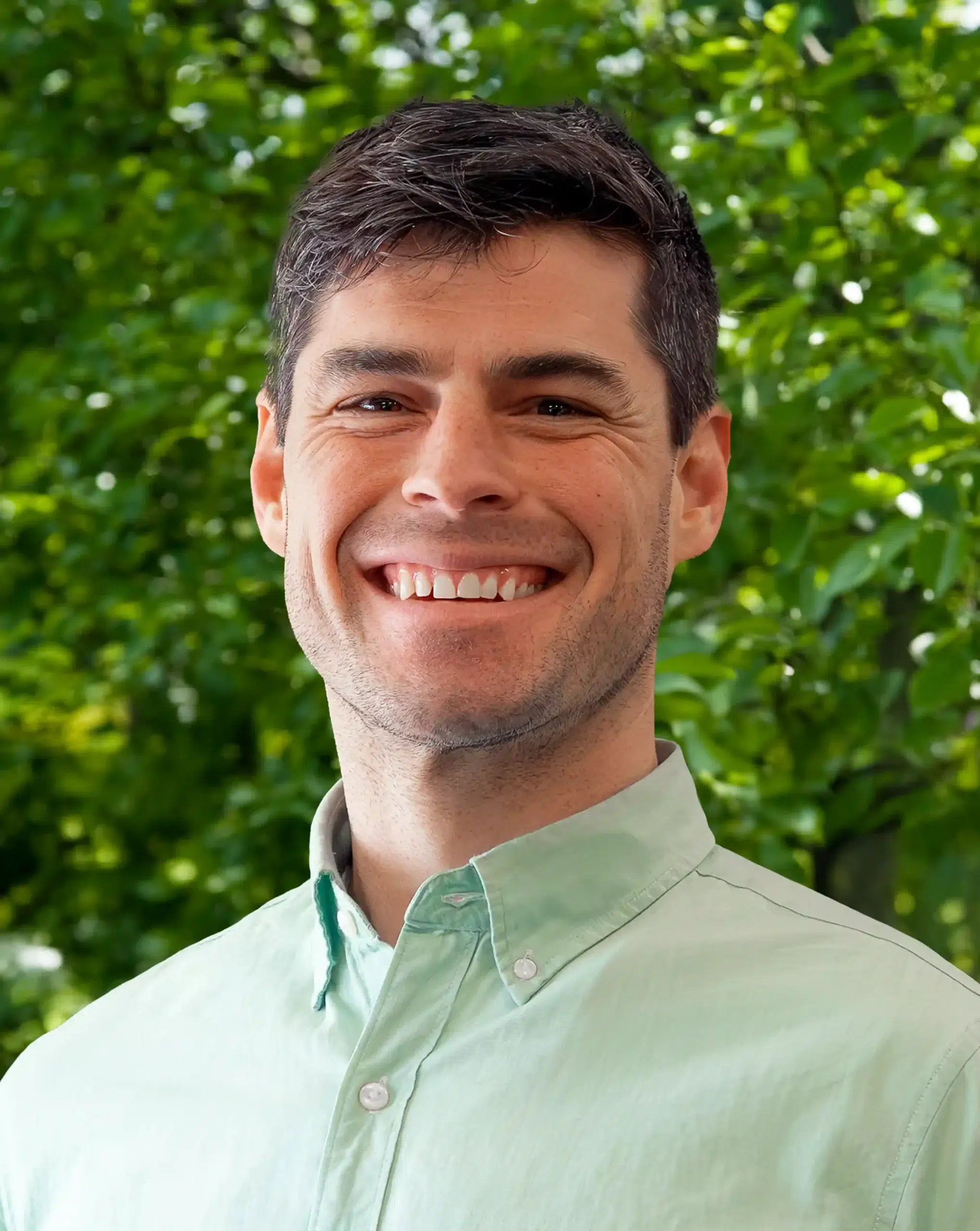 A man with short dark hair smiles while standing outdoors in front of leafy green trees, wearing a light green button-up shirt.