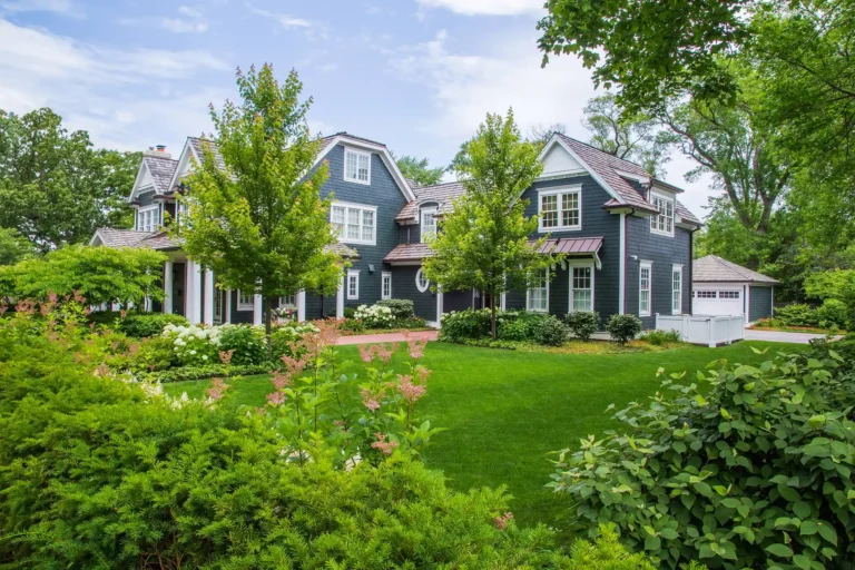 Large blue two-story house with white trim surrounded by lush green lawn, trees, and shrubs on a sunny day.